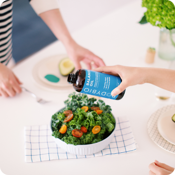 A hand pours BodyBio Balance Oil (Omega 6 + 3), a supplement rich in essential fatty acids, from a blue bottle onto a kale and tomato salad in a white bowl. Two people sit at a white table with plates, half an avocado, and green flowers visible.