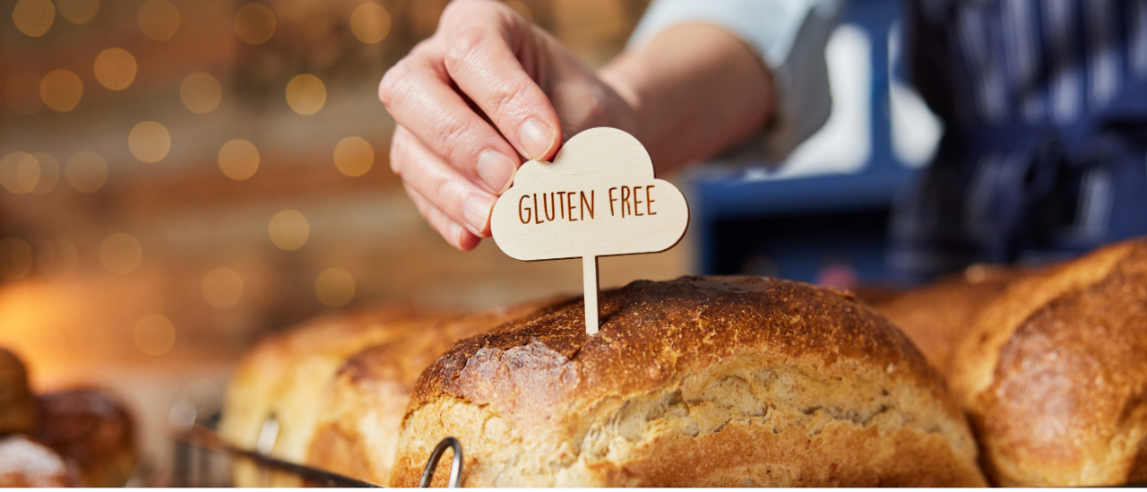 A hand places a wooden "gluten free" sign on a loaf of freshly baked bread with a golden crust at a bakery.