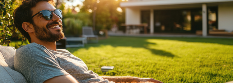 A man with a beard and sunglasses reclines comfortably on outdoor furniture in a sunlit backyard. 