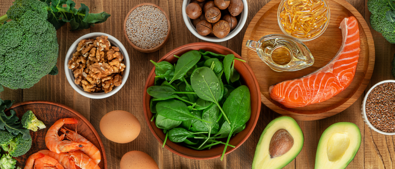 A variety of healthy whole foods including salmon, shrimp, eggs, spinach, avocado, walnuts, broccoli, and oil on a wooden table.