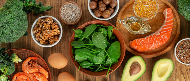 Blog of How Much Fish Oil Should I Be Taking? Experts Answer which have image A variety of healthy whole foods including salmon, shrimp, eggs, spinach, avocado, walnuts, broccoli, and oil on a wooden table.