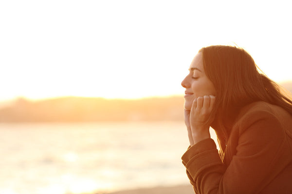 A young woman with long hair and a serene expression rests her chin on her hands while facing the golden sunset at the beach. 