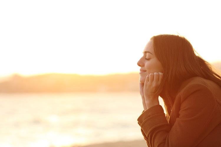 A young woman with long hair and a serene expression rests her chin on her hands while facing the golden sunset at the beach. 