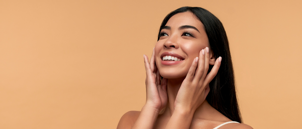 A young woman with radiant, clear skin smiles while touching her face, set against a warm beige background.