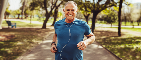 A senior man with a fit physique, wearing a blue athletic shirt and earphones, jogging on a paved path in a green park while smiling. 