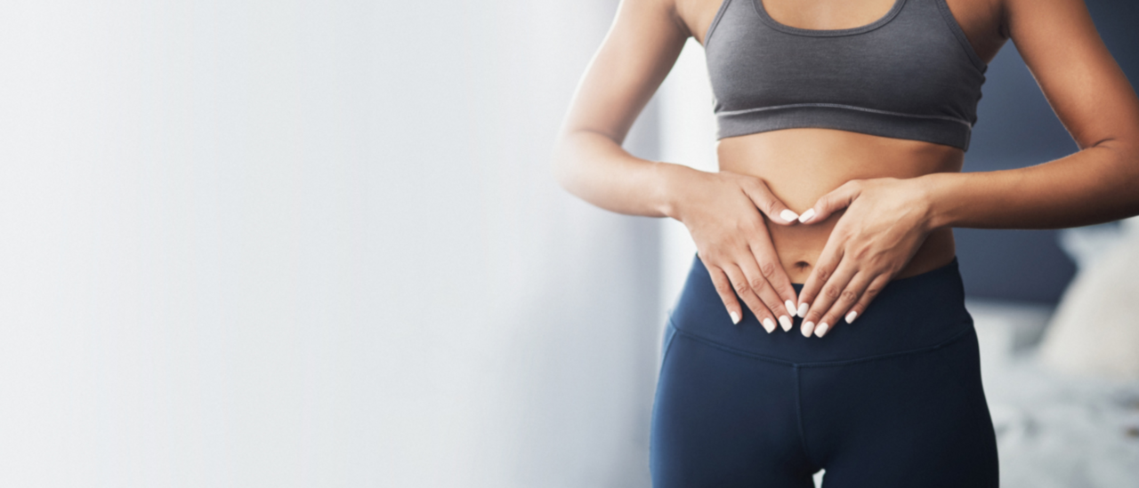 A fit woman in athletic wear forms a heart shape with her hands over her stomach, symbolizing gut health and well-being, with a bright and airy background.