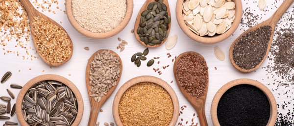 An assortment of seeds, including sunflower, flax, sesame, pumpkin, and chia, displayed in wooden bowls and spoons on a white surface.