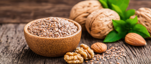 A wooden bowl filled with flaxseeds sits on a rustic wooden surface, surrounded by walnuts, almonds, and fresh green mint leaves, highlighting a selection of healthy, nutrient-dense foods.