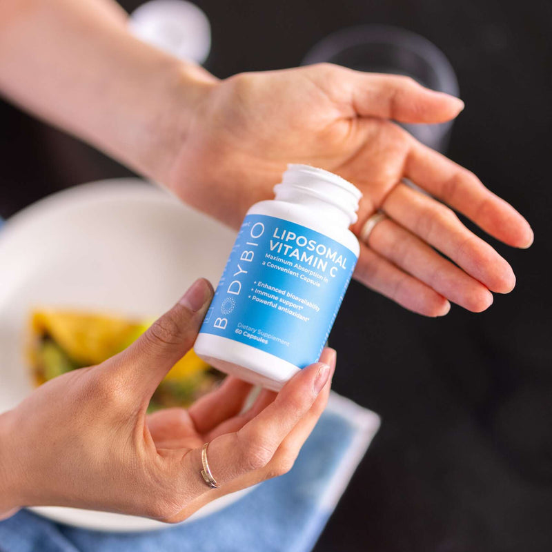 A hand holds a bottle of BodyBio Liposomal Vitamin C. The setting suggests a casual, everyday use environment with a plate of food in the background, emphasizing the supplement's role in daily nutrition and wellness.